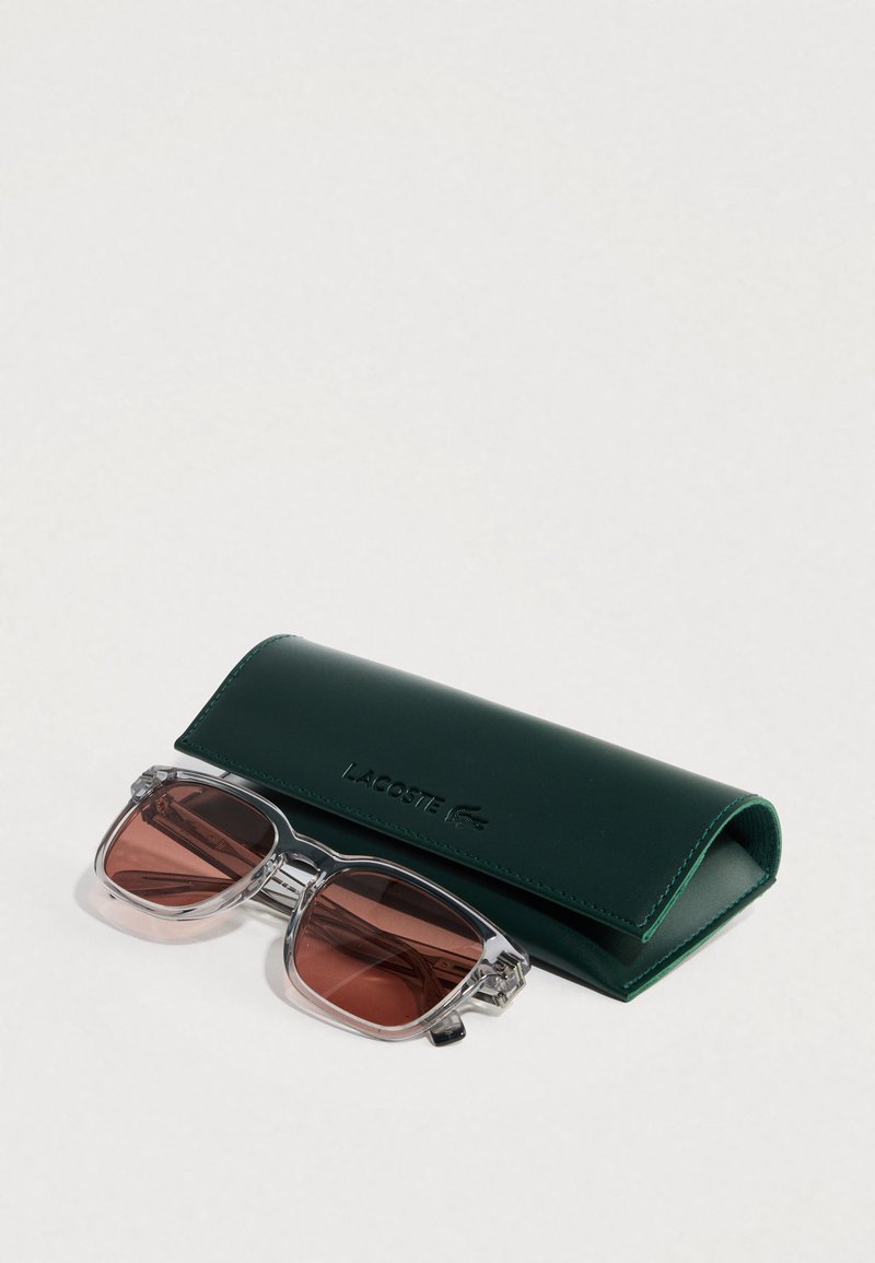 Solbriller med klare stel og brune linser ved siden af et grønt læderetui præget med Lacoste-logoet.