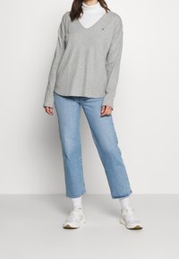 Grå v-hals sweater med lange ærmer, parret med lyseblå straight-leg jeans og hvide sneakers; diskret logo detaljer på sweateren.