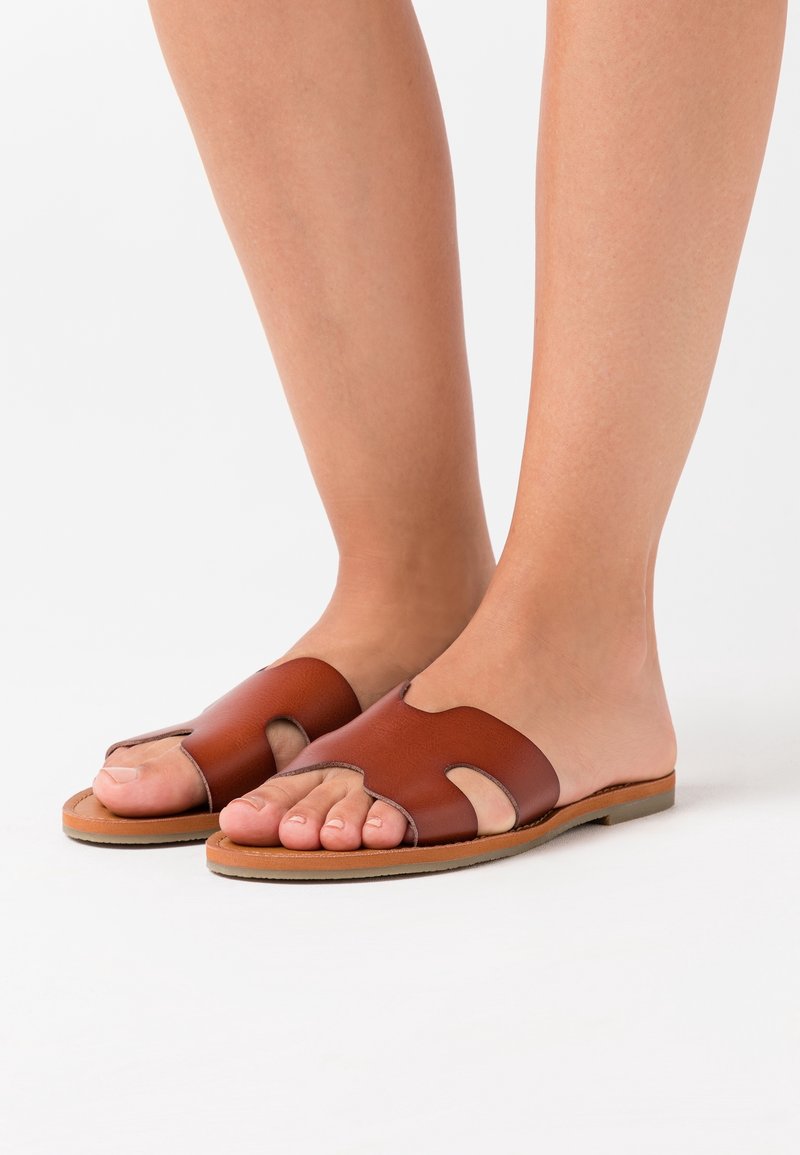 Sandales slides en cuir marron avec des découpes, semelles plates et une texture lisse. Style à bout ouvert mettant en valeur les orteils.
