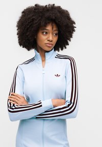 Veste de survêtement à fermeture éclair bleu clair avec des accents rayés noirs et blancs sur les manches. Présente un petit logo sur la poitrine, fabriquée en tissu lisse.