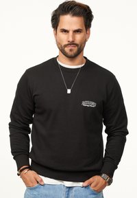 Black sweatshirt with long sleeves, ribbed cuffs, and a crew neckline. Features a small white logo on the left chest and a layered white shirt underneath.