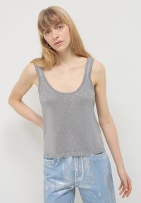 Jeune femme aux cheveux blonds portant un haut sans manches en tricot argenté et un jean métallique bleu clair, se tenant devant un fond blanc uni.