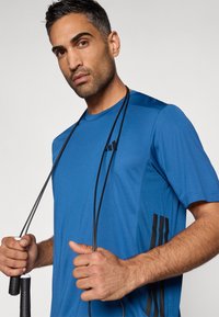 Μπλε αθλητική κοντομάνικη μπλούζα με μαύρες λεπτομέρειες και λογότυπο. Κρατάει σχοινάκι για άλματα, με λεία υφή και δομημένη εφαρμογή.