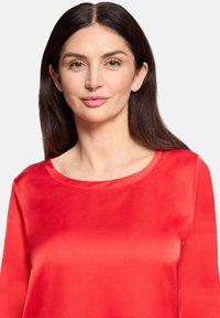 Rød satinbluse med glat tekstur, rund halsudskæring og lange ærmer, med et enkelt design uden mønstre eller accenter.