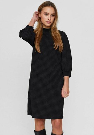 Black dress with a textured, waffle-like pattern, ribbed high neckline, and three-quarter puff sleeves. Knee-length cut with side slits.