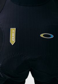 Cicliste negre cu dungi verticale, având o etichetă galbenă și un logo multicolor. Materialul pare elastic, cu detalii de cusătură fine.