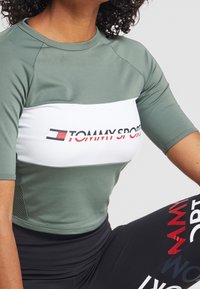 Mujer vistiendo una parte superior atlética verde y blanca de Tommy Sport y leggins negros con texto del logo de Tommy Sport en rojo, blanco y azul.