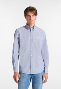 Camicia azzurro chiaro a maniche lunghe, con colletto e logo ricamato sul petto. Abbinata a jeans in denim.
