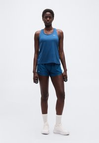 Γυναίκα με μπλε αθλητικό τοπ και σορτς, κρατώντας αλτήρες 1 κιλού, φορά λευκές κάλτσες και αθλητικά παπούτσια, στέκεται μπροστά σε λευκό απλό φόντο.