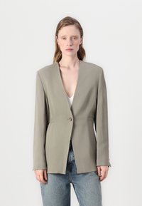 Lys grå tilpasset blazer med dyp V-hals, enkelt knappelukking, strukturerte skuldre og glatt stoff. Båret over en hvit topp.