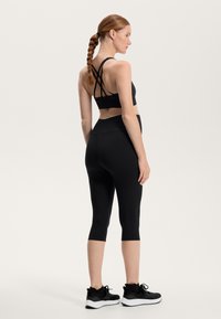 Črn športni kratki top z prepletenimi naramnicami, v kombinaciji z visokim črnim leggingi in črnimi športnimi čevlji z belimi podplati.