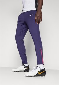 Lila Sporthosen haben einen kontrastierenden pinken Streifen an der Seite und bestehen aus dehnbarem Material. Nike-Logo in Weiß nahe dem Bund.