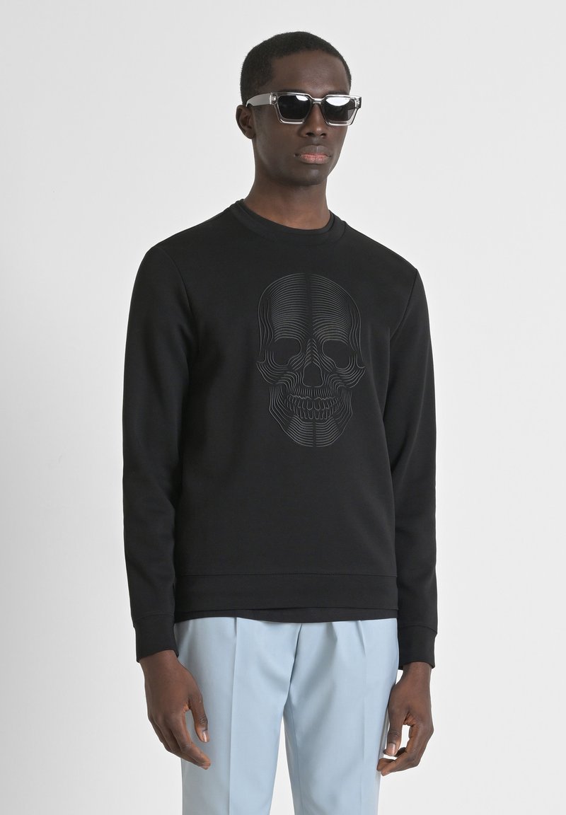 Sudadera negra con un gráfico de calavera texturizado, cuello redondo y puños acanalados. El modelo lleva pantalones azul claro y gafas de sol.