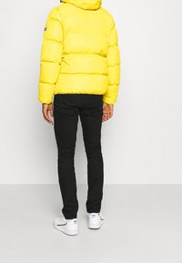 Muž oblečený ve jasně žluté péřové bundě, černých slim-fit kalhotách a bílých teniskách Adidas stojí na jednobarevném pozadí.