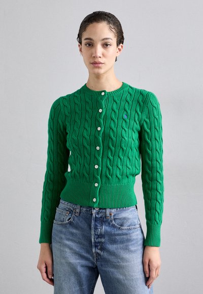 Zelený pletený cardigan s žebrovaným lemem a manžetami. Má kulatý výstřih a zapínání na knoflíky s bílými knoflíky, v kombinaci s modrými džíny.