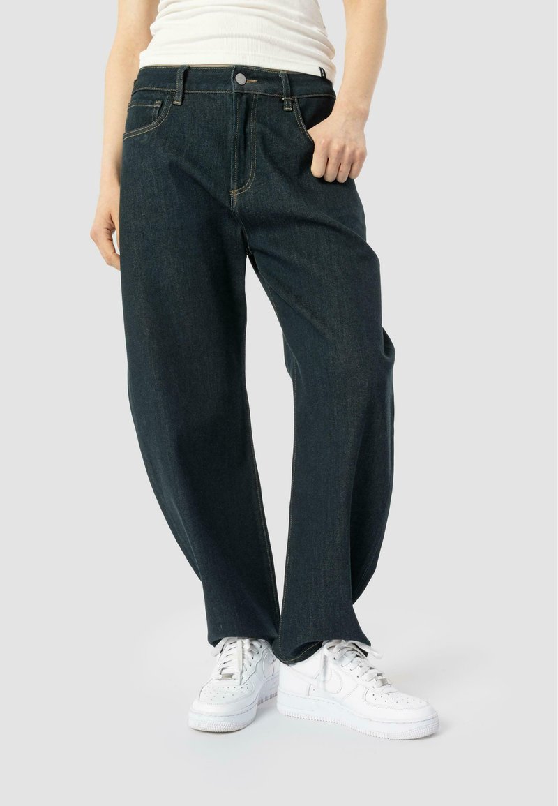 Dark blue, loose-fitting denim pants with contrast stitching, five pockets, and a button fly, paired with white sneakers.