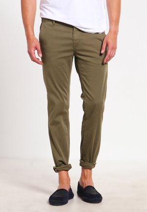 Mann trägt olivgrüne hochgekrempelte Hose, weißes Hemd und dunkelblaue Slipper, steht vor einem schlichten hellen Hintergrund.