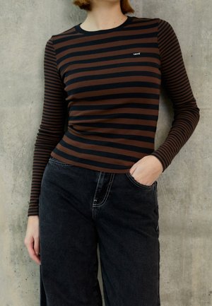 Personne portant une chemise à manches longues rayée marron et noire avec le logo Levi's et un jean noir taille haute, debout contre un mur en béton.