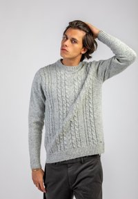Maglione lavorato a maglia grigio chiaro con motivo a treccia, dotato di un collo rotondo e polsini e orlo a coste. Indossato con pantaloni scuri, dalla texture liscia.