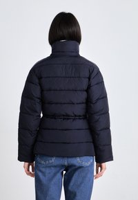 Námornícky puffer kabát s vysokým golierom, prešívanými detailmi a priliehavým pásom. Nosený cez svetlomodré džínsy, s hladkou, izolovanou textúrou.