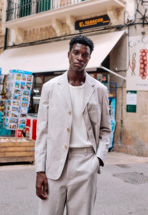 Muž v svetlobéžovom obleku a bielej košeli stojí na ulici, v pozadí sú pohľadnice a obchod s tabakom.