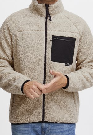 Personne portant une veste polaire beige texturée avec une fermeture éclair noire et une poche poitrine zippée noire, mains jointes devant, sur fond blanc.