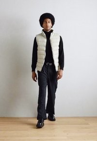 Padded cream vest over a black turtleneck, paired with striped wide-leg trousers and black shoes, worn by a model with an Afro hairstyle.