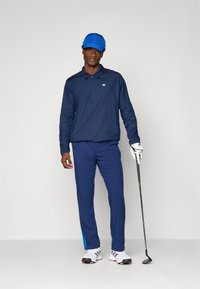 Homem em boné azul, polo azul-marinho e calças azuis, segurando um taco de golfe, usando luva de golfe branca e sapatos de golfe brancos com riscas pretas.