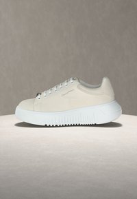 Κρεμ αθλητικό παπούτσι με λευκή χοντρή σόλα, σχεδιασμό με κορδόνια και διακριτική σήμανση στο πλάι, τοποθετημένο σε στρογγυλή επιφάνεια.