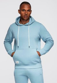 Homme portant un sweat à capuche bleu clair et un pantalon de jogging assorti, mains dans la poche du sweat, regardant droit devant sur un fond uni.