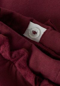 Bordowy materiał z połączeniem gładkiej i puszystej tekstury. Posiada przezroczysty guzik oraz biały metek z kwiatowym wzorem.