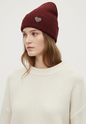 Bordeauxrote, gerippte Strickmütze mit Leopardenherz-Akzent. Modell trägt einen cremefarbenen, strukturierten Pullover.