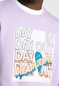 Lila sweatshirt med en central grafisk design med fet text "DAG UT OCH DAG IN" och "ANDAS DJUPARE", accentuerad med blå och orange färger.