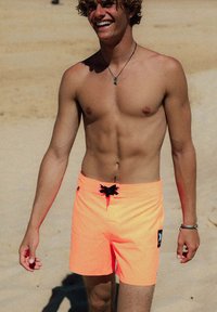 Neonoranje zwembroek met zwarte trekkoord en label. Gladde stoftextuur, boven-de-knie pasvorm, geschikt voor strandkleding.