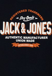 Mornarski majica s izrazitim bijelim i narančastim printom. Tekst uključuje "JACK & JONES", "AUTENTIČNI PROIZVOĐAČ" i "OSNOVANO 1990." Mekana textura.