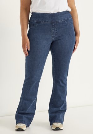 Kvinde iført mørkeblå bootcut-jeans og beige sneakers står mod en hvid baggrund.