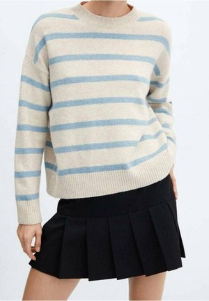 Person iført en cremefarvet sweater med lyseblå vandrette striber og et sort plisseret skørt, der står mod en ensfarvet baggrund.