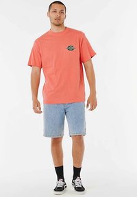Korallenfarbenes Baumwoll-T-Shirt mit kurzen Ärmeln und einem grünen Motiv auf der Brust, kombiniert mit hellen Jeansshorts und schwarzen Turnschuhen.