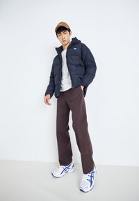 Veste matelassée bleu marine avec fermeture éclair, pantalon ample marron et baskets blanches avec des accents bleus. Le modèle porte une casquette beige. Fond uni.