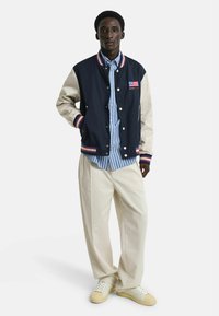 Giacca varsity blu navy e beige con accenti a righe a costine, indossata sopra una camicia a righe blu e bianca, abbinata a pantaloni larghi color crema e sneakers.