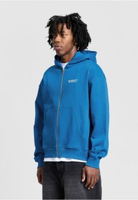 Blauwe rits-hoodie van zachte stof, met een voorzak, een capuchon met trekkoorden en een logo op de borst. Geribbelde mouwen en onderrand.
