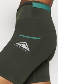 Close-up van donker groene Nike Trail fietsbroek met teal accenten en reflecterend logo op de heup en bovenbeen van een persoon.