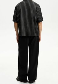 Černá pruhovaná košile s krátkým rukávem a uvolněným střihem, v kombinaci s volnými černými kalhotami, obě s hladkým materiálem a minimalistickým designem.