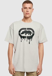 Tricou din bumbac crem cu un grafic mare negru al unui rinocer care picură, însoțit de un logo circular deasupra. Mâneci scurte, croială lejeră.