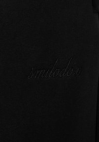 Zwarte stof met subtiele geborduurde tekst "smilodon" in bijpassend zwarte draad, met zichtbare textuur en stiksel details.