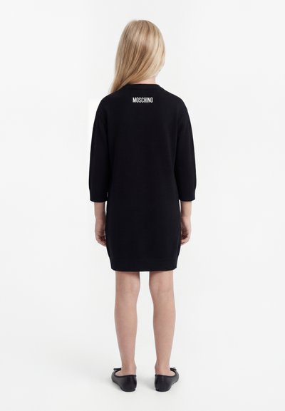 Copil cu păr blond lung, purtând o rochie-pulover neagră Moschino și balerini negri, stând cu spatele spre un fundal alb.