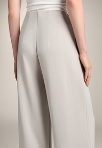Ljusgrå vida byxor med en slät textur och subtila sömnadsdetaljer, visade från baksidan för att framhäva den skräddarsydda passformen och draperingen.