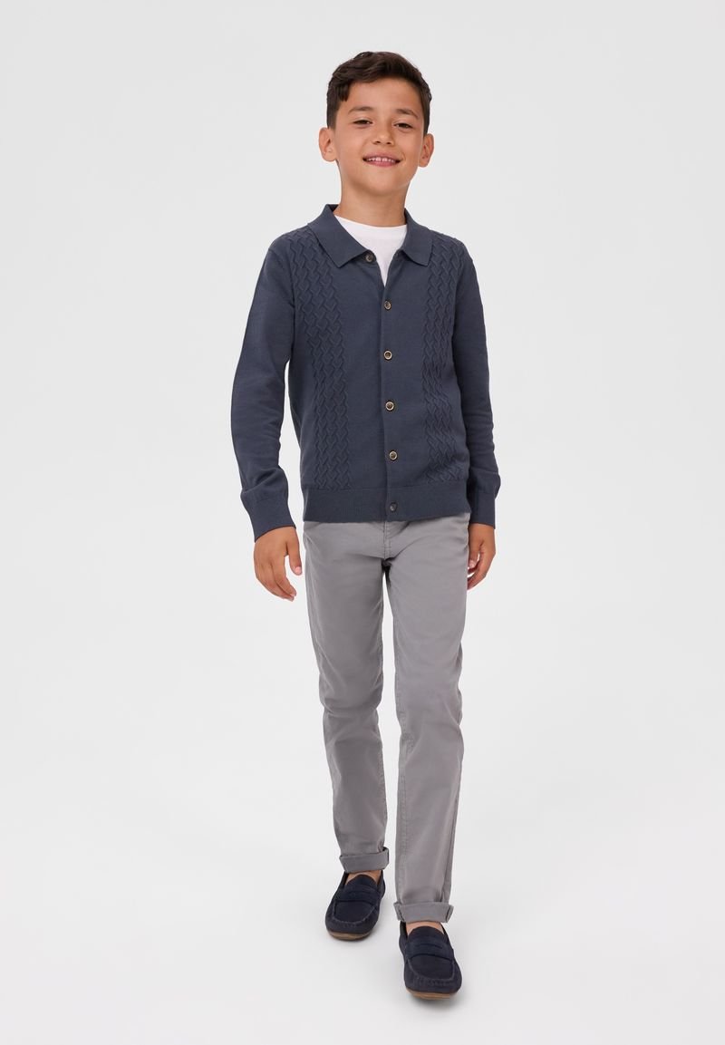 Garçon marchant, portant un cardigan bleu marine boutonné, un maillot blanc, un pantalon gris clair, et des mocassins bleu marine, sur fond blanc uni.