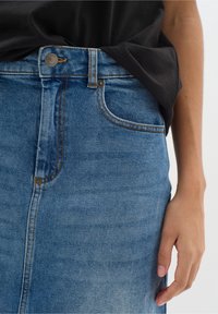 Jeanskjol i ljusblått med figursydd midja, synliga sömmar och en framfick, kombinerad med en svart t-shirt. Har en knappstängning.