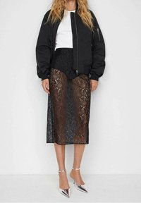 Blouson aviateur noir sur une jupe midi en dentelle noire, avec des sections transparentes et des motifs complexes, associé à des escarpins argentés à bout pointu.